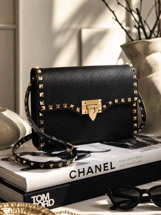 A black pebbled leather handbag with gold-tone studs and a gold clasp, styled on top of books with a Chanel magazine and sunglasses nearby.
