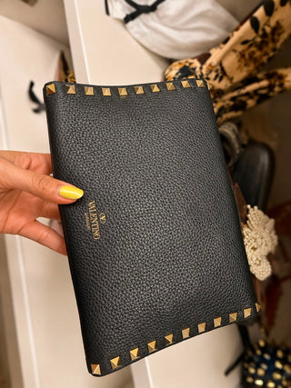 A black pebbled leather wallet with gold pyramid studs and a gold Valentino logo, held by a hand with yellow nail polish against a cluttered closet background.