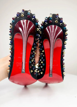 A pair of high-heeled stiletto shoes with bright red patent soles and black uppers heavily embellished with multicolored rhinestones and gold-toned spikes, held against a plain light background.