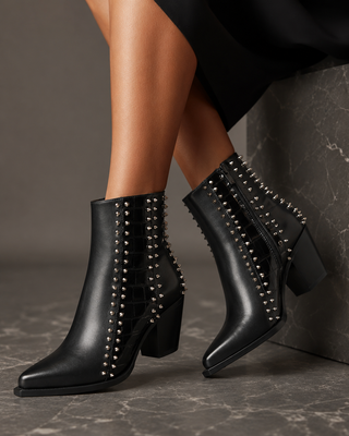 Black leather ankle boots with silver studded side panels and block heels, shown on a model's feet against a dark gray stone background.
