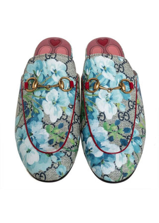 A pair of Gucci GG Supreme canvas loafers featuring a light blue and white floral print, gold-tone horsebit detailing, and red piping, shown against a plain white background.