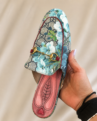 A hand holds a Gucci slip-on loafer-style shoe featuring a blue and white floral print over the brand's signature GG monogram canvas, with a gold horsebit detail and a pink insole.