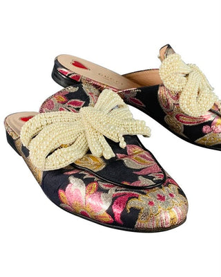 A pair of Gucci slip-on mule flats featuring a black satin floral brocade base with pink, gold, and green metallic embroidery, accented by large cream pearl-beaded bow details across the top.