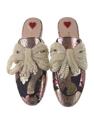 A pair of Gucci flat mule sandals featuring a black and pink paisley brocade base, adorned with large cream pearl-beaded bows across the top and a red heart logo on the insole.