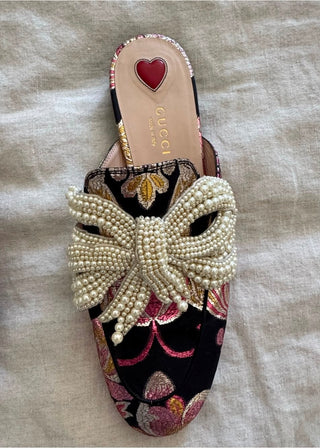 A Gucci slip-on mule shoe featuring a large pearl-beaded bow on a black velvet base with pink and gold floral embroidery, laid flat on a beige linen background.