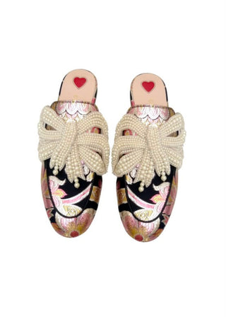 A pair of embellished mule flats featuring a black base covered in pink and gold floral embroidery, adorned with large cream pearl-beaded bow straps and a small red heart on the insole.