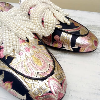 A pair of ornate traditional Chinese brocade shoes decorated with white pearl bead tassels and laces, displayed on a distressed white wooden surface.