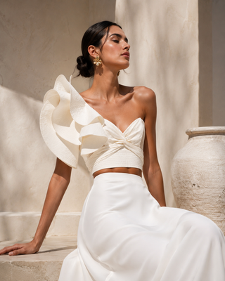 Woman wearing a white bridal gown with ruffled top and flowing skirt against a neutral background.