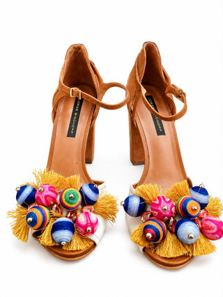 A pair of tan leather high-heeled sandals with ankle straps, decorated with bright multicolored spherical beads and yellow tassels on the open toes, shown against a plain white background.
