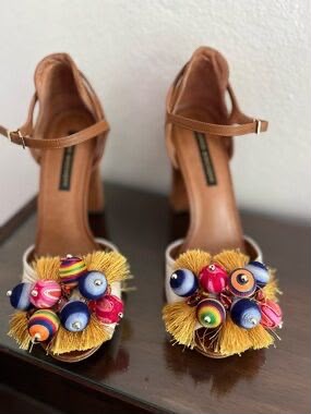 A pair of tan block-heel sandals with ankle straps, decorated with colorful glass-like beads and yellow tassels on the toes, displayed on a dark wooden surface against a light wall.