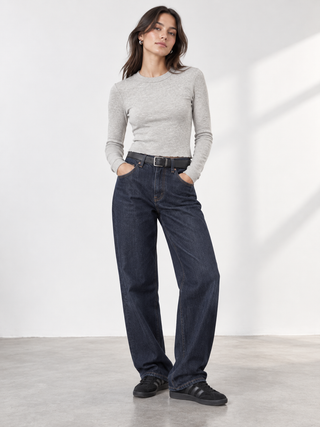 A woman modeling a light gray long-sleeve knit top and dark blue wide-leg jeans with a black belt, standing in a bright studio with soft window shadows.