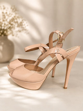 A pair of nude beige platform high-heeled sandals with ankle straps and gold-tone hardware, displayed on a warm beige surface with soft shadows and a blurred vase of flowers in the background.