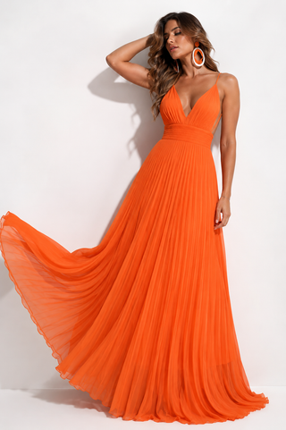 A woman modeling a bright orange sleeveless maxi dress with a deep V-neckline and a flowing pleated skirt that spreads out on the left side, set against a plain white studio background.