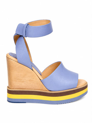 A light blue leather wedge sandal with an ankle strap, a thick natural wood wedge heel, and a multi-colored platform sole featuring yellow, brown, and blue stripes, shown against a plain white background.