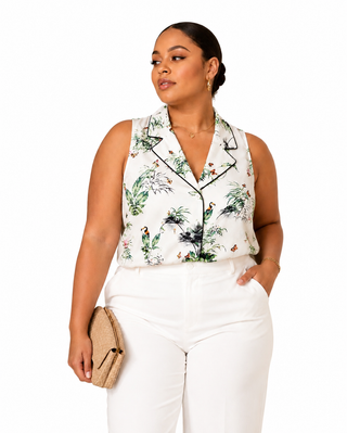 A woman modeling a sleeveless white button-up blouse with a tropical bird and plant print, styled with white trousers and a beige clutch bag against a plain white background.