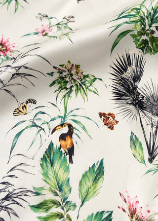 A close-up of a tropical fabric print featuring a toucan, butterflies, palm fronds, and green leaves on a cream background.