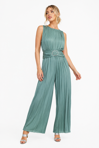 A woman modeling a sleeveless pleated jumpsuit in a muted sage green color, featuring a defined waist with a silver ring buckle belt and wide-leg pants, set against a plain white studio background.
