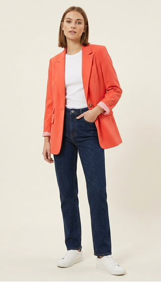 Woman wearing an orange blazer, white shirt, blue jeans, and white sneakers on a beige background