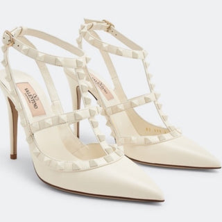 A pair of Valentino pointed-toe stiletto pumps in off-white leather, decorated with signature pyramid studs along the straps and ankle ties, shown on a light gray background.
