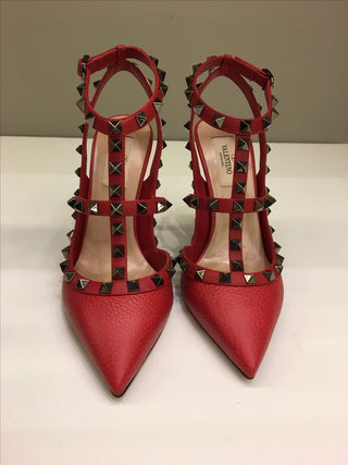A pair of red pointed-toe high heels with multiple straps, black and silver pyramid studs, and a visible Valentino insole.