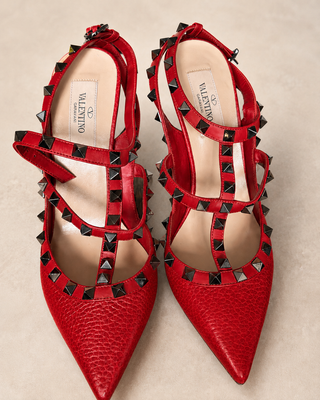 A pair of red pointed-toe slingback heels by Valentino, featuring textured leather, crisscross ankle straps, and black pyramid studs around the edges, photographed on a neutral beige background.