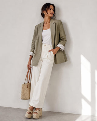 Woman wearing a green blazer, white top, and white pants, holding a straw bag against a white wall.